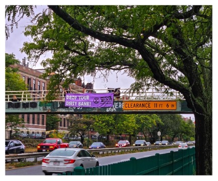 A photograph of a foot bridge over an urban highway. A tree grows in the foreground. From the footbridge hangs a purple banner with the words DROP YOUR DIRTY BANK!