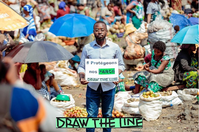 A man in jeans and a collared shirt standing in a large crowd of people holds a small sign that reads Protegeons nos PARCS DRAW THE LINE.