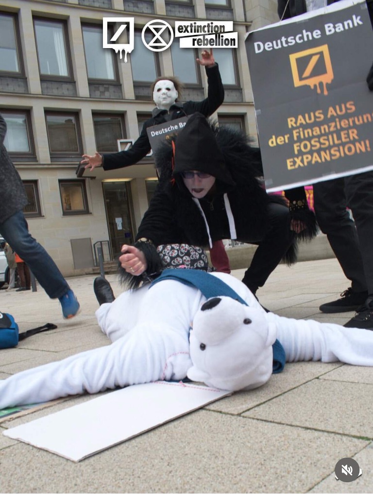 A photo of a street-theater scene in an outdoor urban space. Someone in a polar bear suit is laying on the ground, apparently being stabbed in the chest by a character in black fur. Another performer is holding a sign that says "Deutsche Bank raus aus der finanzieruns fossiler expansion"!