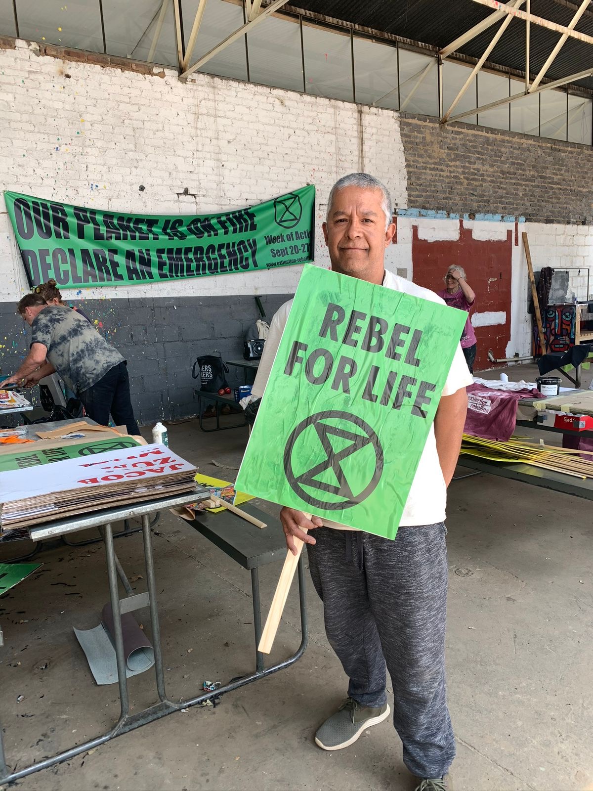 A photo of a man in a warehouse-like space holding a green sign that says REBEL FOR LIFE