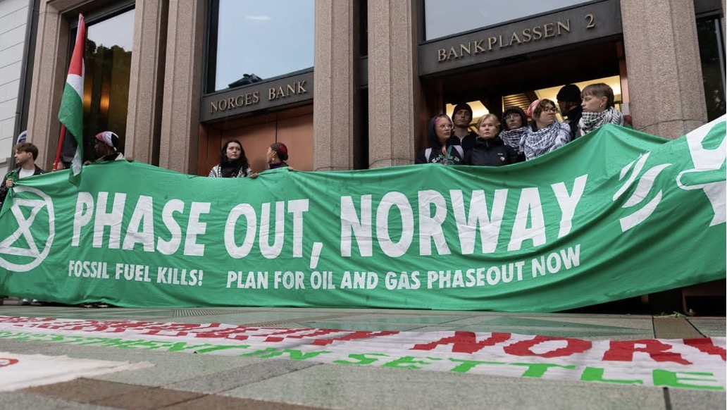 A photo of a small group of people holding a large banner in front of a building. Signs on the building read "Norges Bank" and "bankplassen 2". The banner is green with white lettering and reads "phase out, Norway" and, in smaller lettering, "fossil fuel kills! Plan for oil and gas phaseout now". One person also holds a flag, though it's hanging limp and so too little of its pattern is visible to recognize. Two other banners lay on the ground in front of the people, but these are at the wrong angle to read.